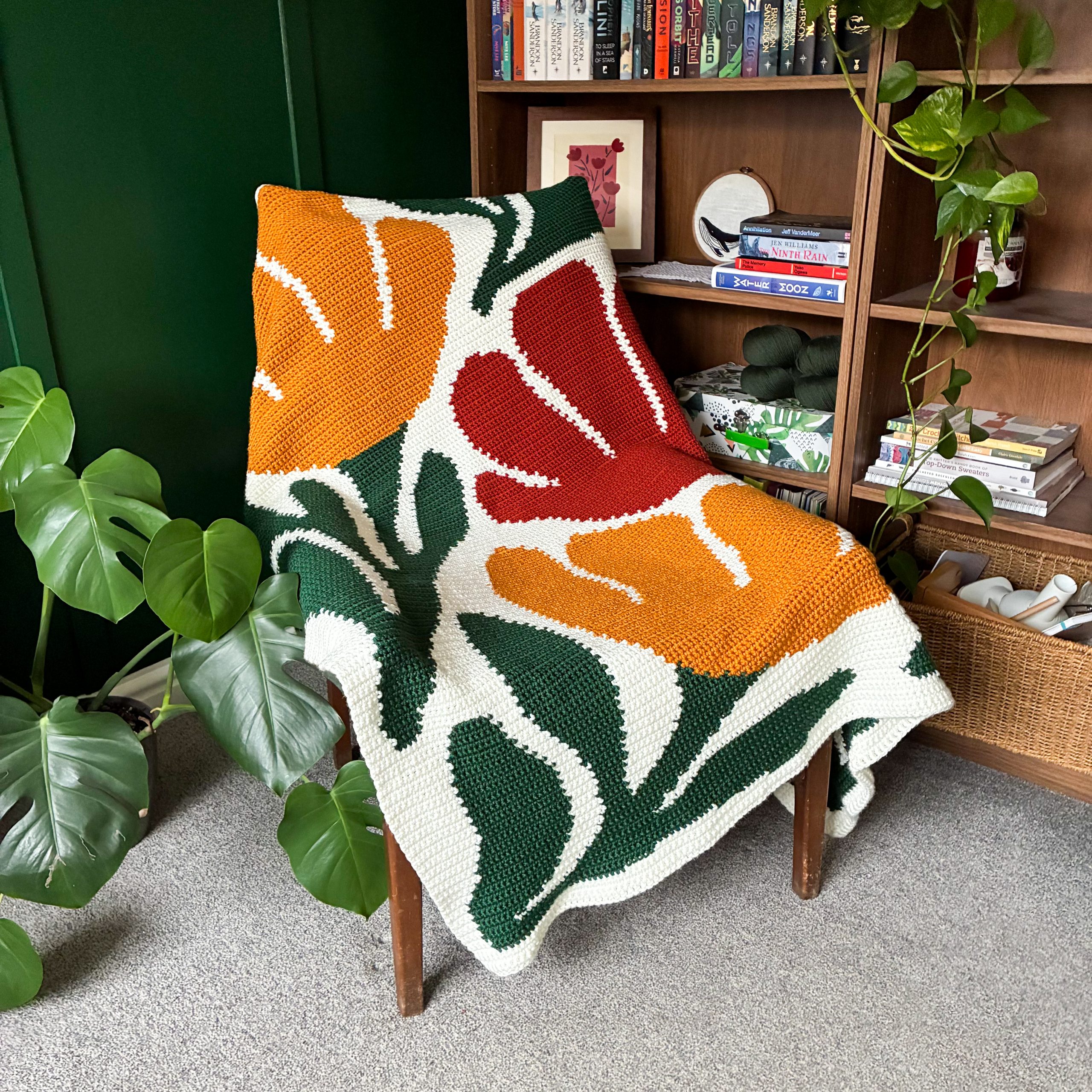 Bloom By Bloom Blanket Chair Bloom By Bloom crochet flower blanket draped over a chair in a cozy reading corner, featuring a colorful floral graphgan design in orange, red, dark green, and cream.