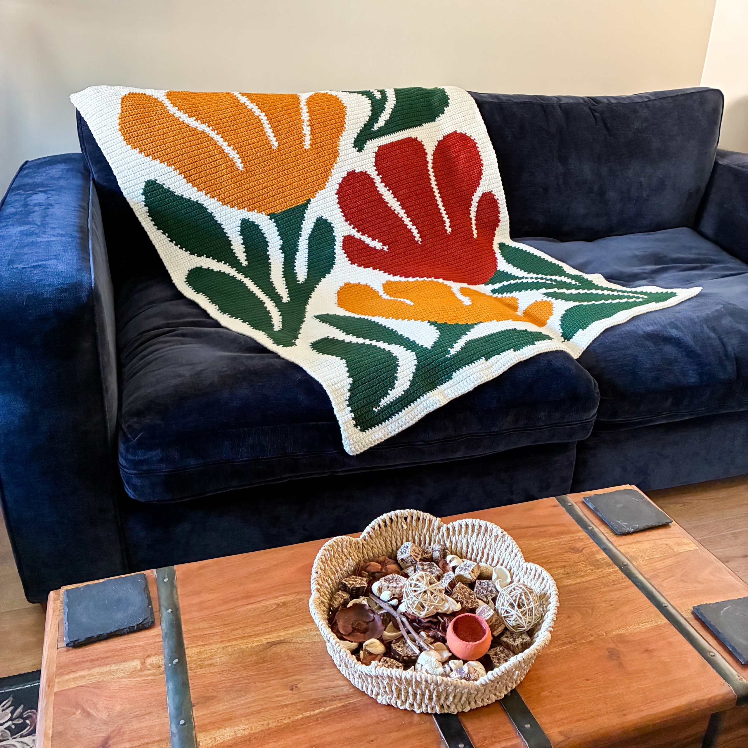 Bloom By Bloom Blanket Sofa + Coffee Table Bloom By Bloom floral crochet graphgan blanket draped across a navy blue sofa, featuring three oversized flowers in red and orange with dark green leaves on a cream background.