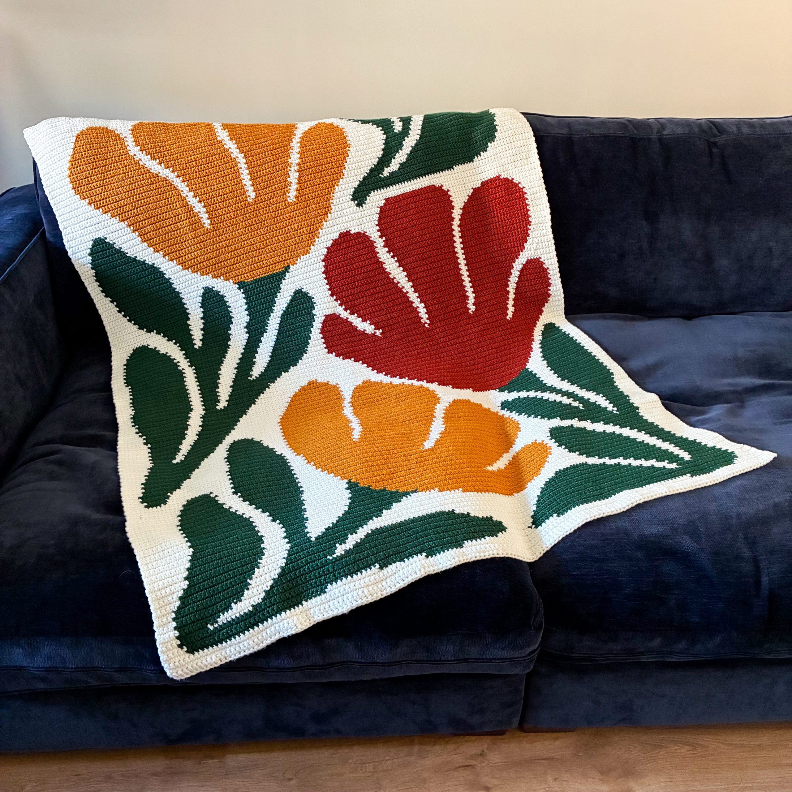 Handmade Bloom By Bloom crochet blanket displayed on a navy blue sofa, showing the complete floral graphgan design with bold red and orange blooms and green foliage.
