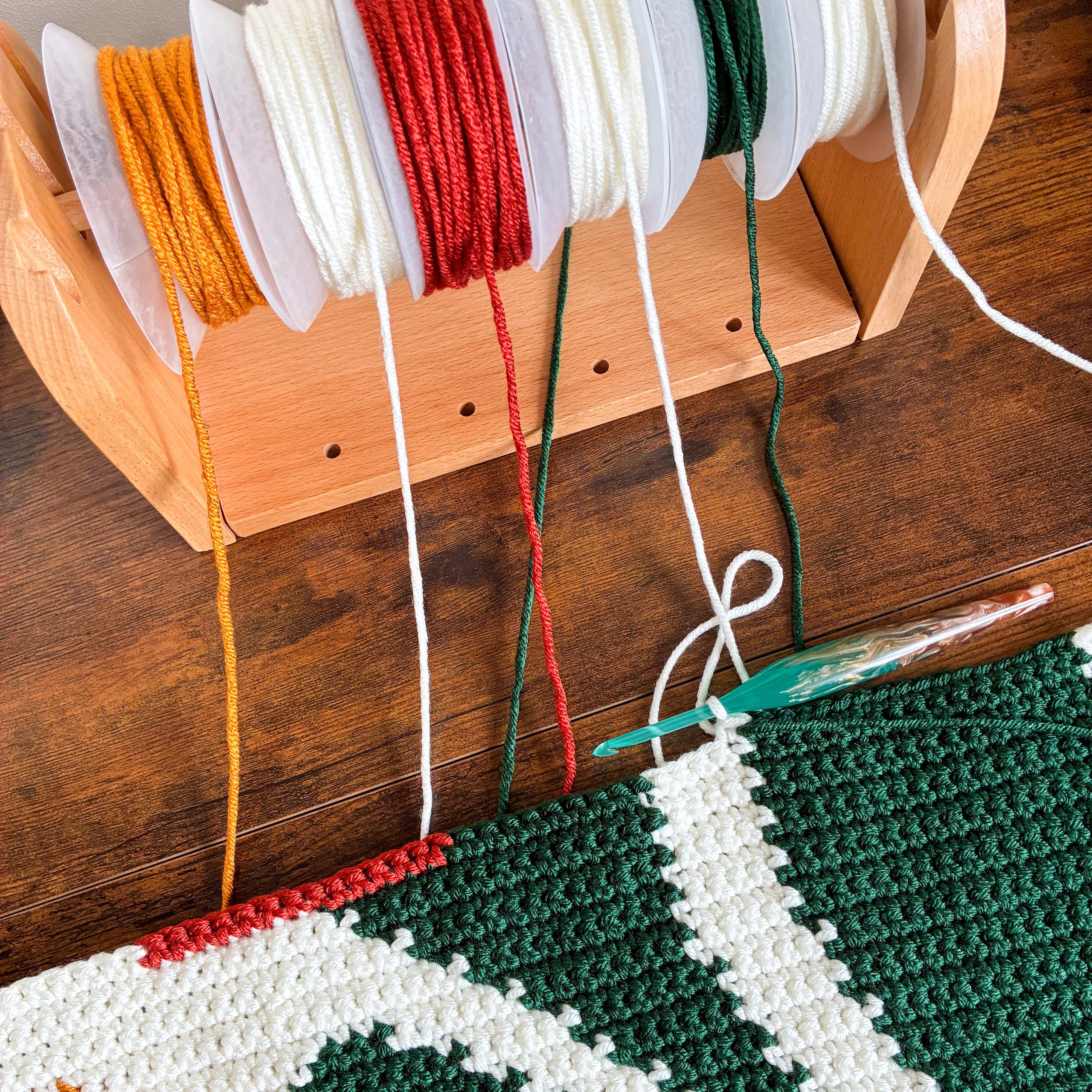 Bloom By Bloom Blanket WIP Work in progress on the Bloom By Bloom crochet graphgan blanket, with orange, cream, red, and dark green yarn wound onto bobbins on a wooden yarn holder.