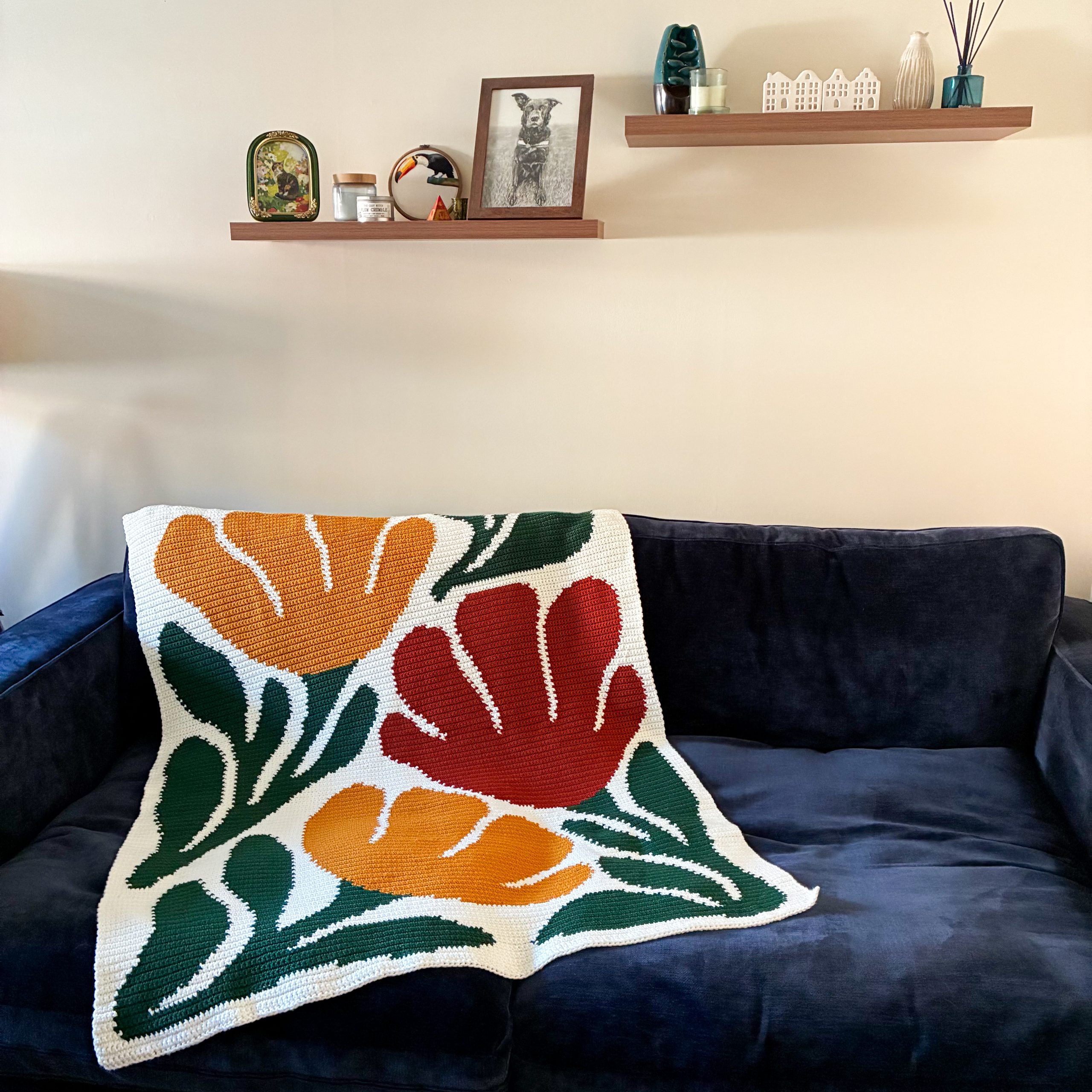 Bloom By Bloom Blanket Wide Angle Wide view of the Bloom By Bloom crochet graphgan blanket draped over a navy blue sofa in a living room, featuring oversized floral motifs in warm orange, red, and dark green.