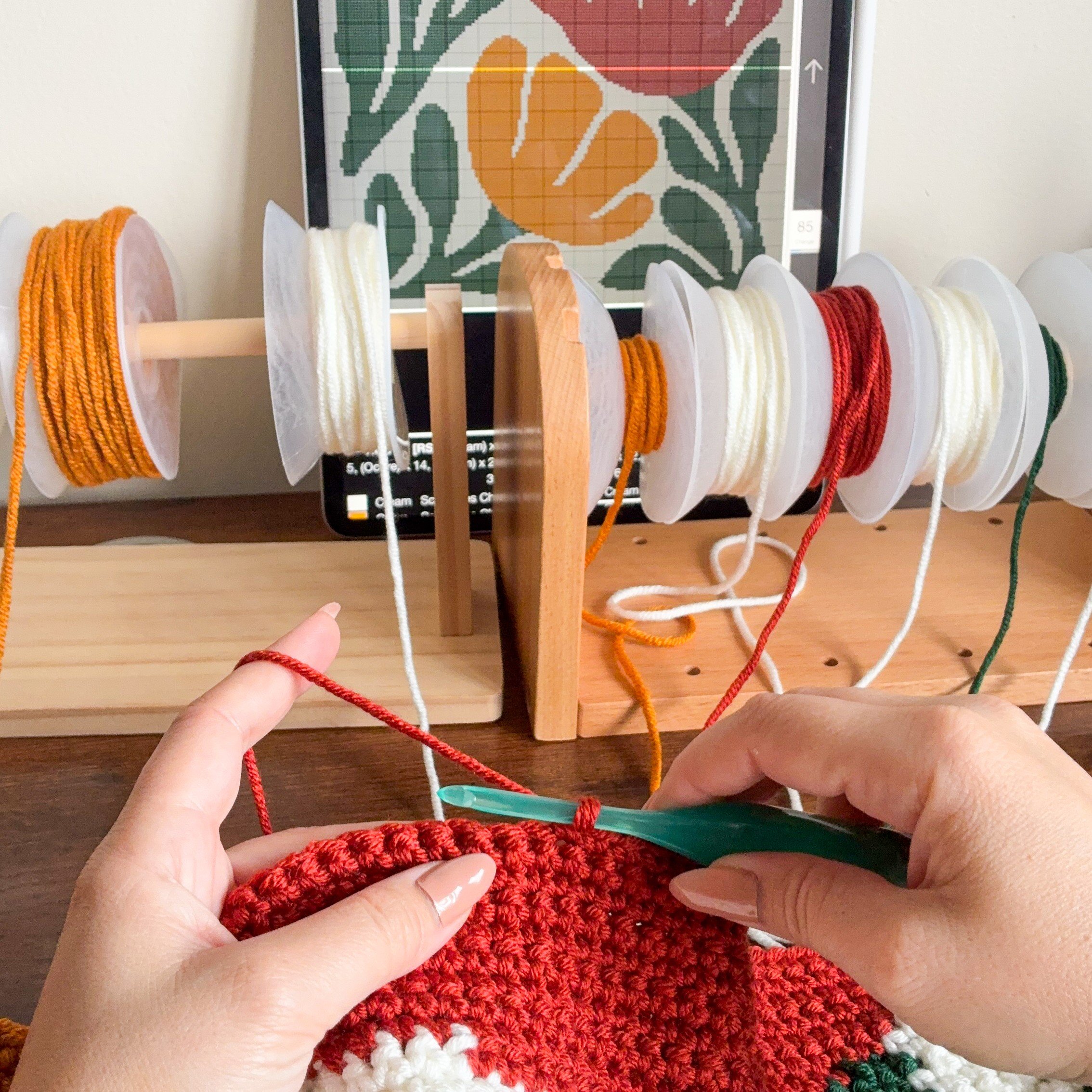 Bloom By Bloom Blanket Work in progress Hands crocheting the Bloom By Bloom floral graphgan blanket using red yarn, with yarn bobbins on wooden holders and a digital colorwork chart displayed on a tablet.