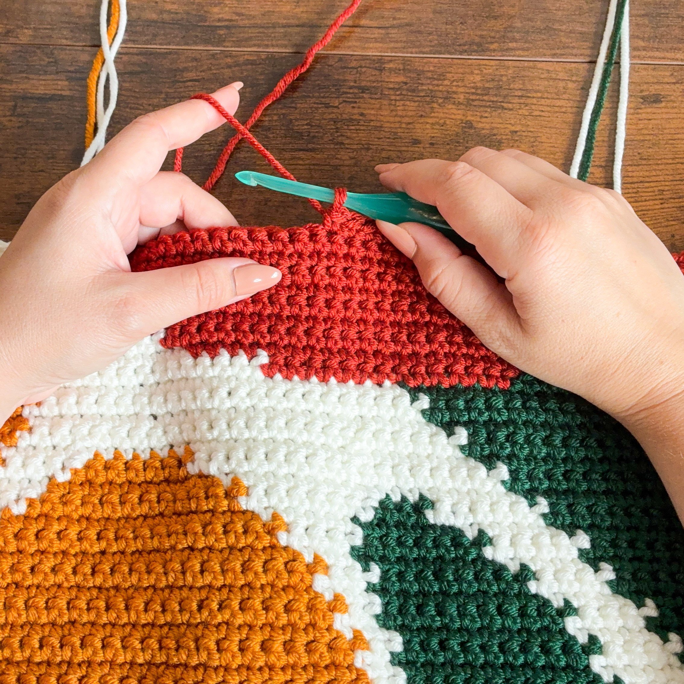 Bloom By Bloom Blanket crocheting Close-up of hands crocheting the Bloom By Bloom graphgan blanket with red yarn and a green crochet hook, showing the colorwork stitches forming the floral design.