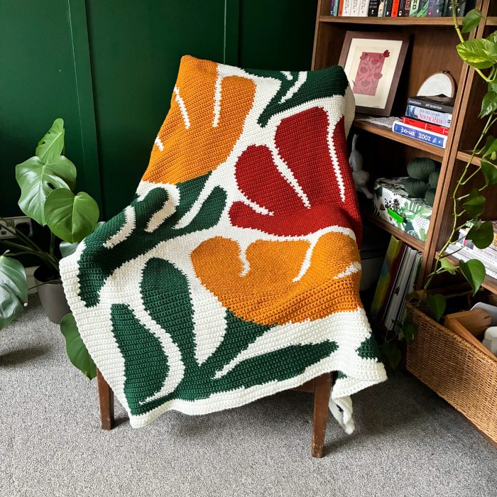 Bloom By Bloom crochet graphgan blanket draped over a wooden chair beside a bookshelf, featuring oversized orange and red flowers, dark green leaves, and a cream background.