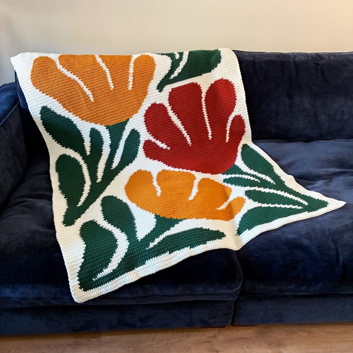 Handmade Bloom By Bloom crochet blanket displayed on a navy blue sofa, showing the complete floral graphgan design with bold red and orange blooms and green foliage.